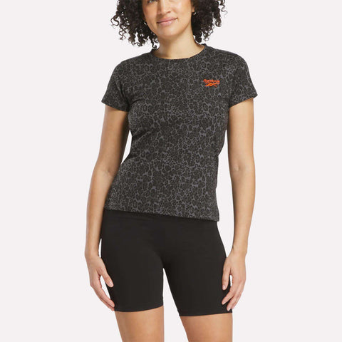A woman with curly hair wears a black Reebok Women's Leopard Print Baby T-Shirt, featuring leopard print and an orange logo, paired with black bike shorts against a plain white background.