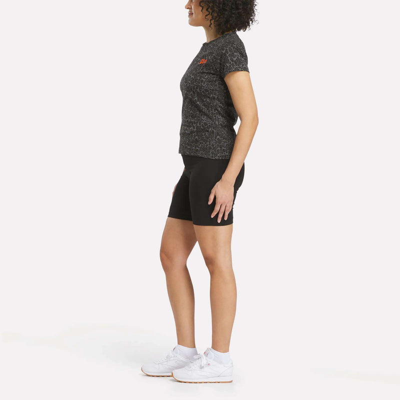 A woman stands in profile against a white background wearing the Reebok Women's Leopard Print Baby T-Shirt, black biker shorts, and white sneakers.