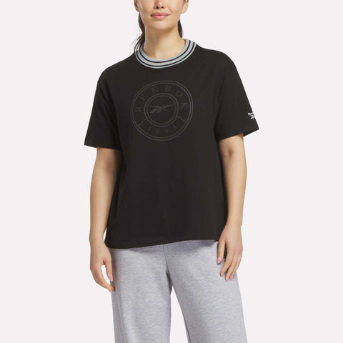 A woman wears a black Reebok Women's Lawn Club T-Shirt featuring a circular logo on the front, paired with grey sweatpants and a striped crew neckline, against a plain light background.