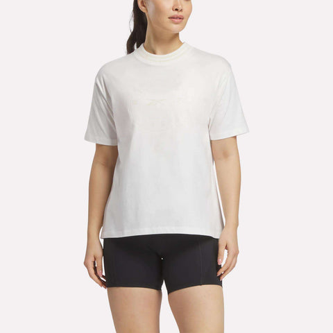 A woman in a loose Reebok Women's Lawn Club T-Shirt and black fitted shorts stands against a plain background, looking slightly to the side with her arms relaxed by her sides.