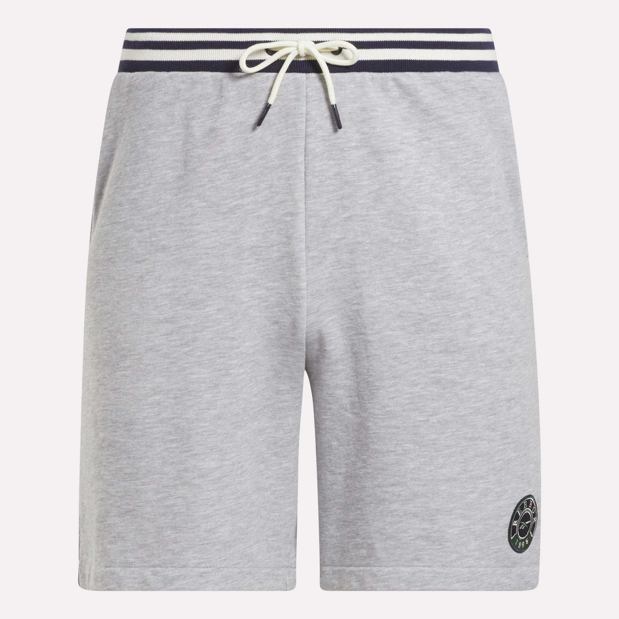 The Reebok Men's Lawn Club Shorts are light gray with a navy and white striped elastic waistband, white drawstring, side pockets, and a small circular logo on the lower left leg. Made from a comfortable cotton blend.