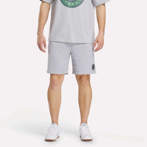 A person faces forward wearing Reebok Men's Lawn Club Shorts in a cotton blend, a light gray t-shirt, and white athletic sneakers against a plain, light-colored background.