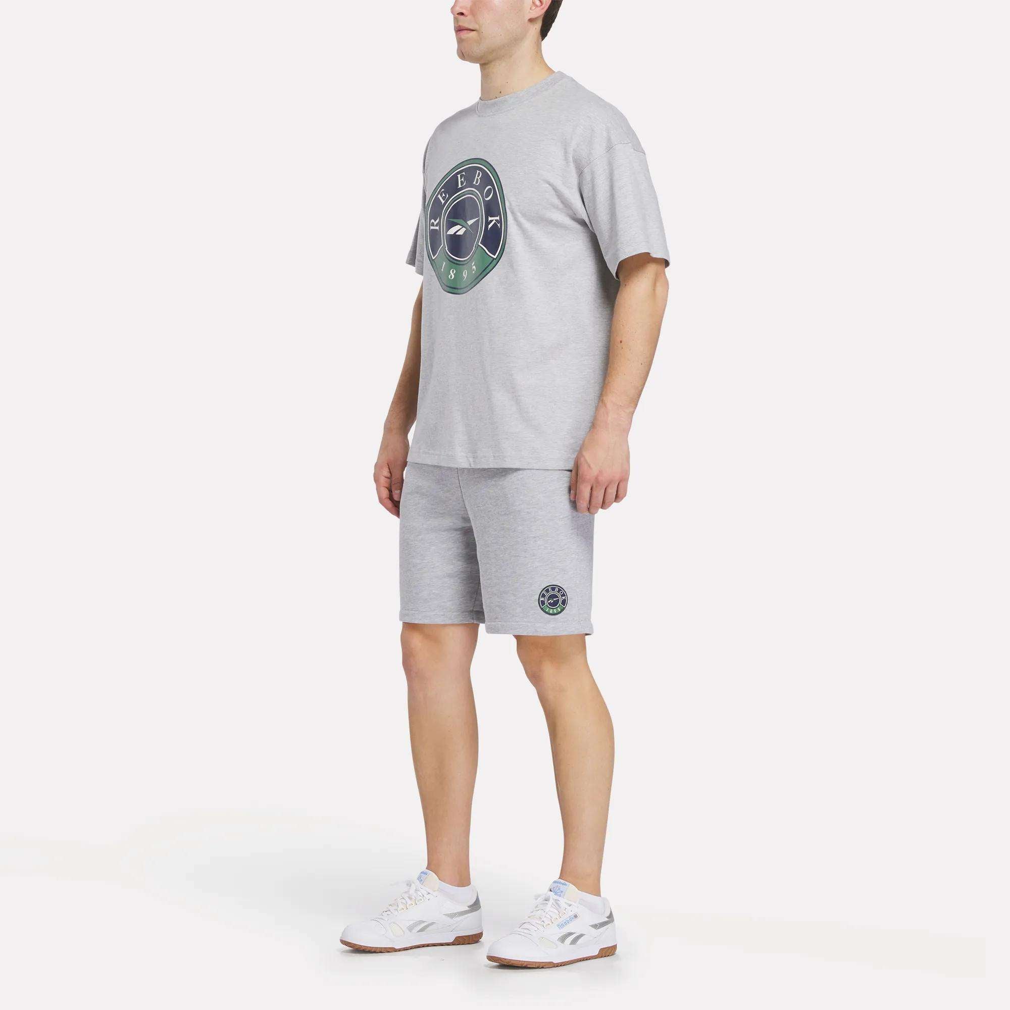 A person wearing a light grey T-shirt with a circular logo, Reebok Men's Lawn Club Shorts, and white sneakers stands against a plain white background.