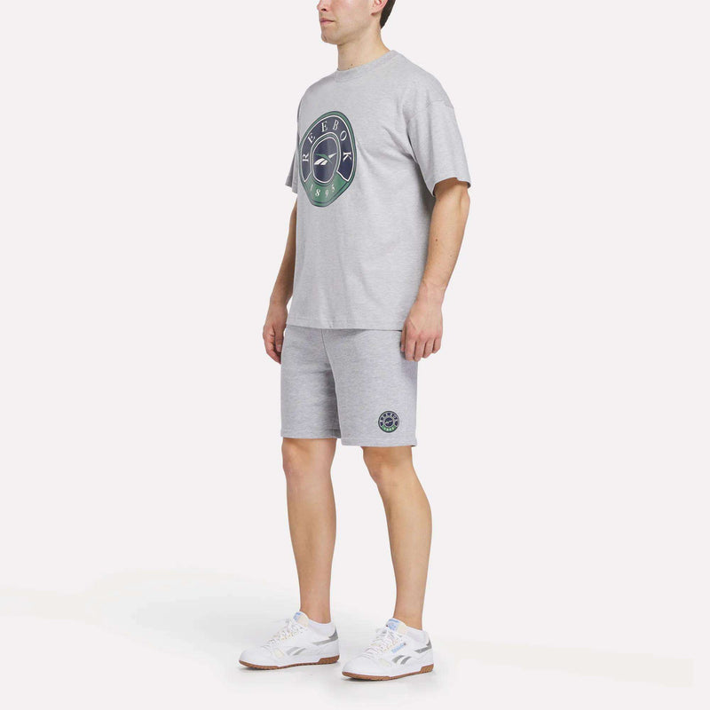 A person wearing a light grey T-shirt with a circular logo, Reebok Men's Lawn Club Shorts, and white sneakers stands against a plain white background.