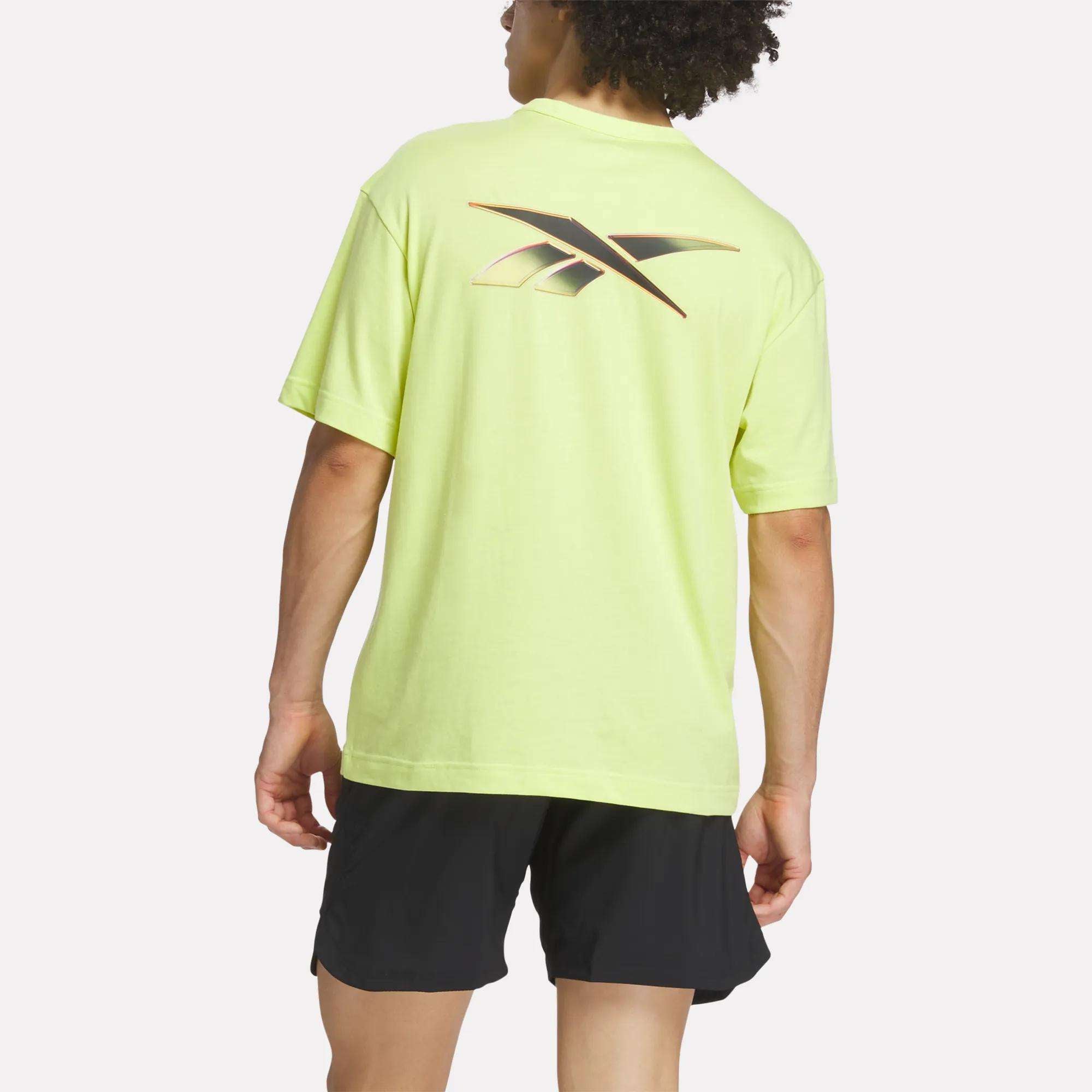 A person wearing a light yellow Reebok Men's Athlete Speed Logo T-Shirt with a large back logo and black shorts, standing and facing away from the camera.