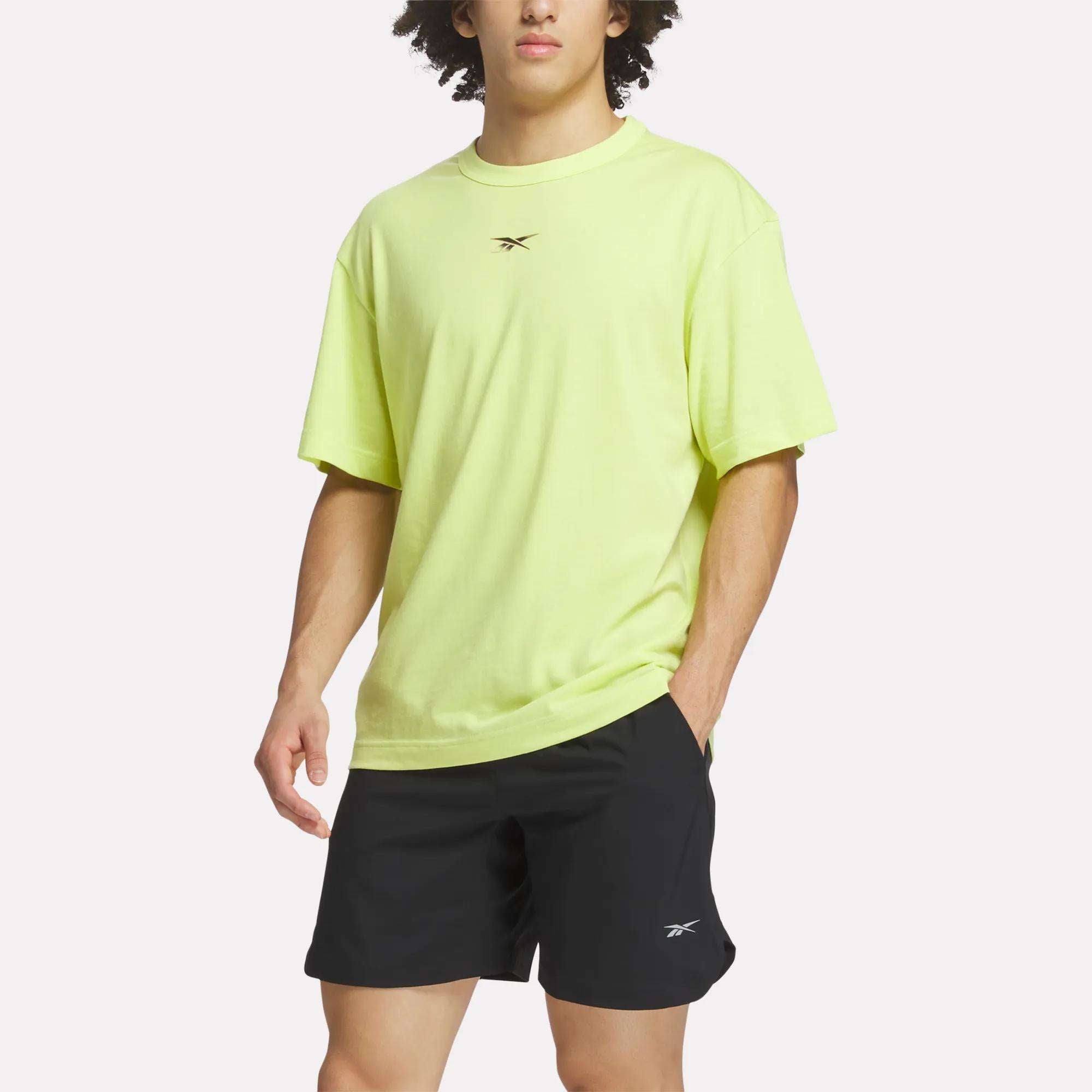 A person wearing a loose-fitting, light yellow Men's Athlete Speed Logo T-Shirt by Reebok and black Reebok shorts stands with one hand in their pocket against a plain, light background.