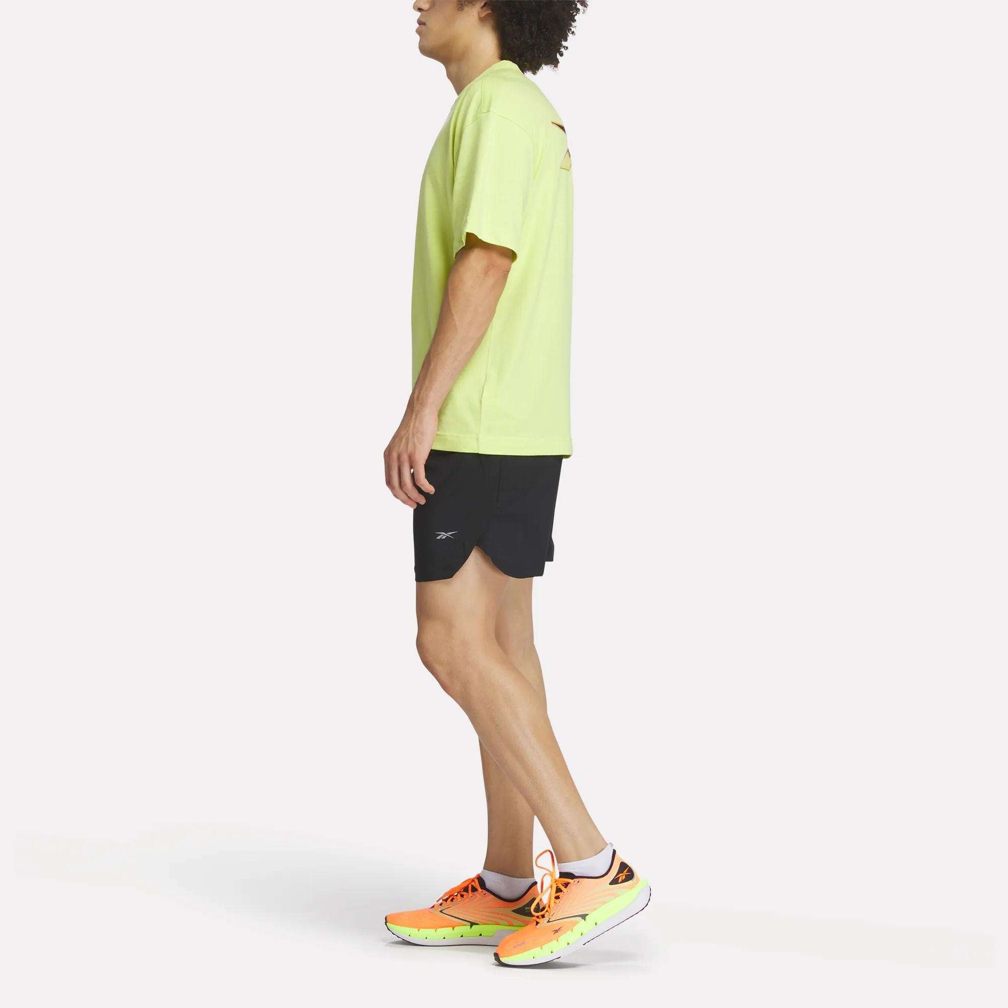 A person stands sideways wearing a light green Reebok Men's Athlete Speed Logo T-Shirt, black shorts, and bright yellow-orange running shoes with white soles against a plain white background.