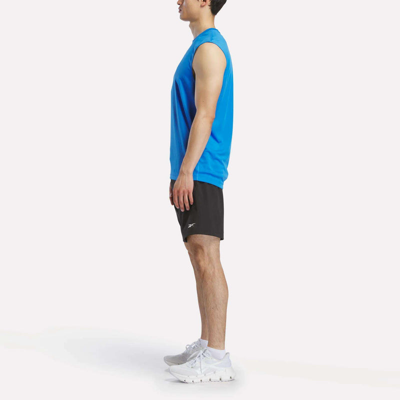 A man stands in profile facing left, wearing a Reebok Men's Sleeveless Tech Training T-Shirt, black shorts, and white athletic shoes against a plain white background.