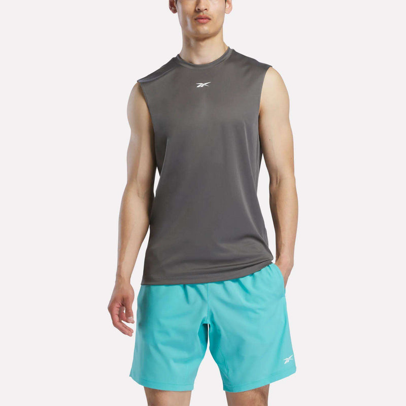 A man wears a Reebok Men's Sleeveless Tech Training T-Shirt in dark gray with light blue shorts, standing facing forward against a plain light background.