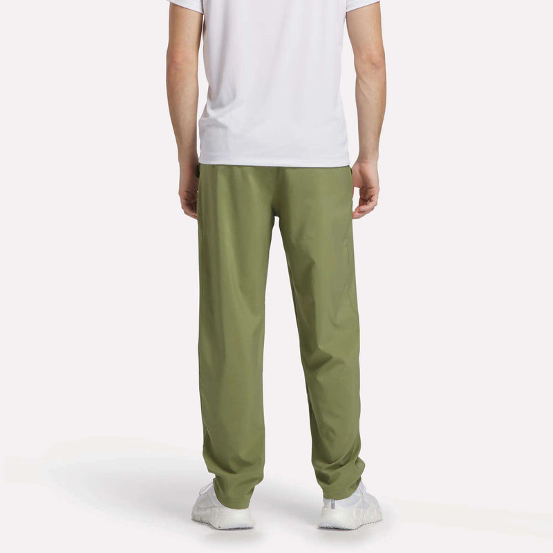 A person wearing a white short-sleeve shirt, Reebok Men's Train Woven Unlined Pants in light green, and white sneakers stands facing away from the camera against a plain white background.