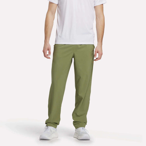 A man is shown from shoulders down wearing Reebok Men's Train Woven Unlined Pants in light green, paired with a white short-sleeve shirt and white sneakers, set against a plain white background.
