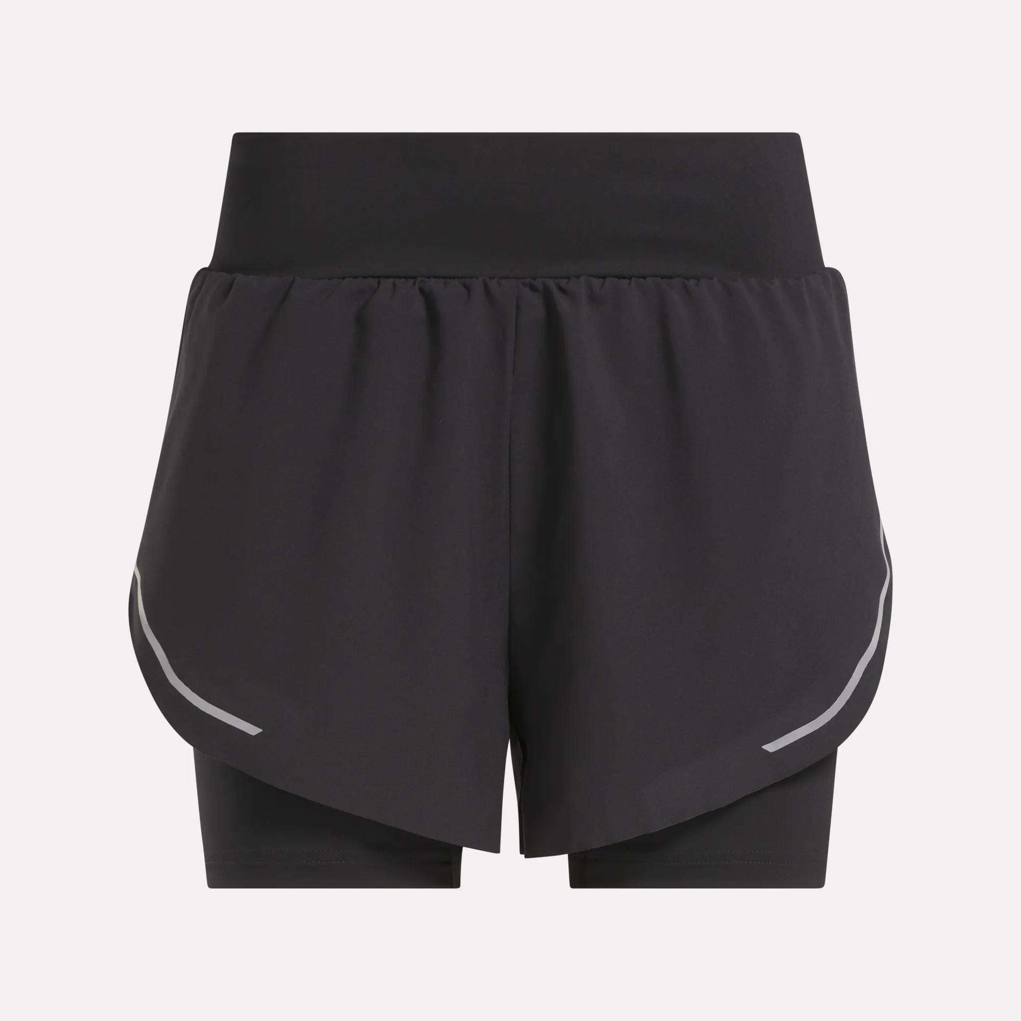 The Reebok Women's Lux Metallic 2-in-1 Running Shorts 3" are black, high-waisted shorts with an overlapping front layer, reflective curved side stripes, and fitted inner shorts. Shown against a plain light gray background.