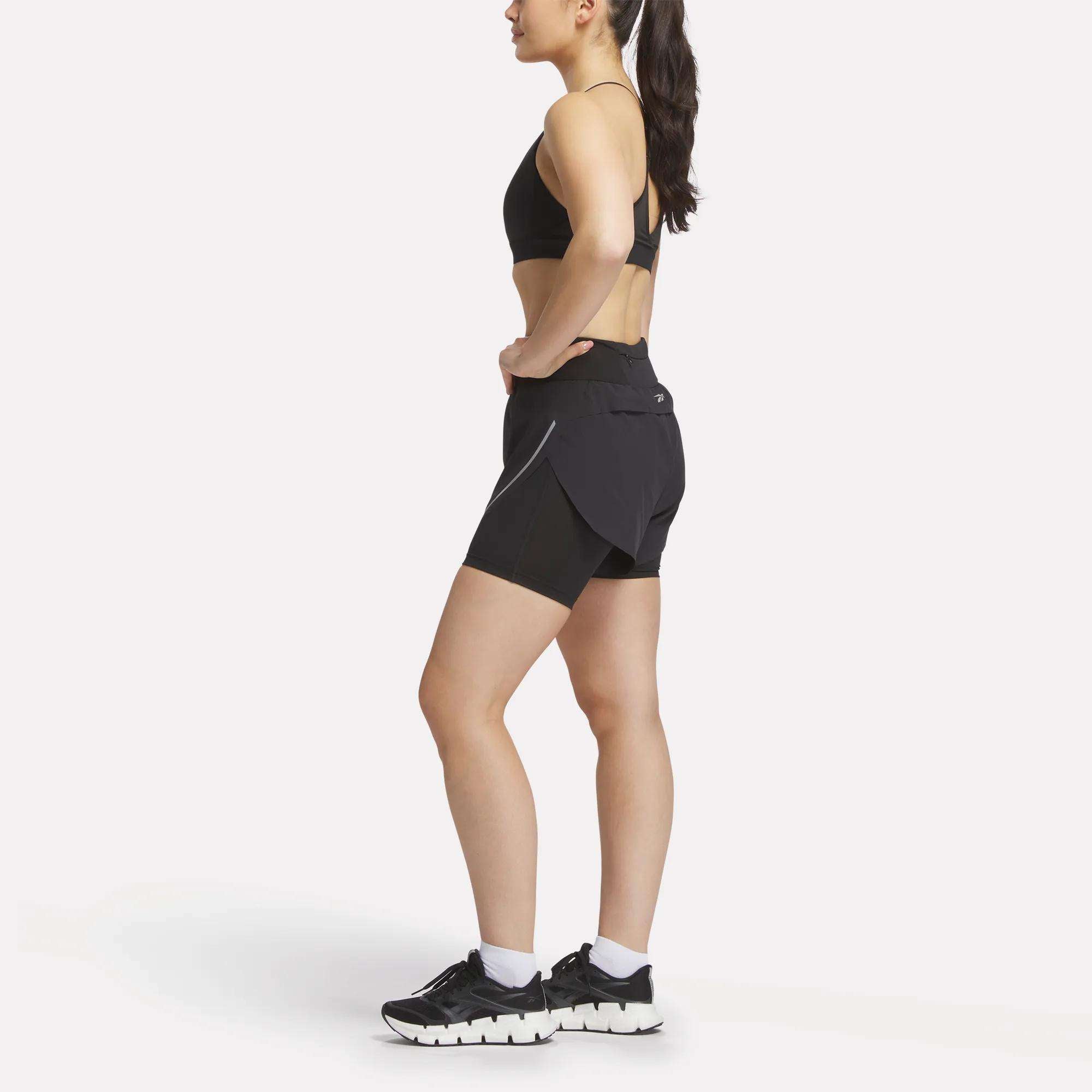 A woman in profile wears a black sports bra, Reebok Women's Lux Metallic 2-in-1 Running Shorts 3", white socks, and black athletic shoes against a plain white background.