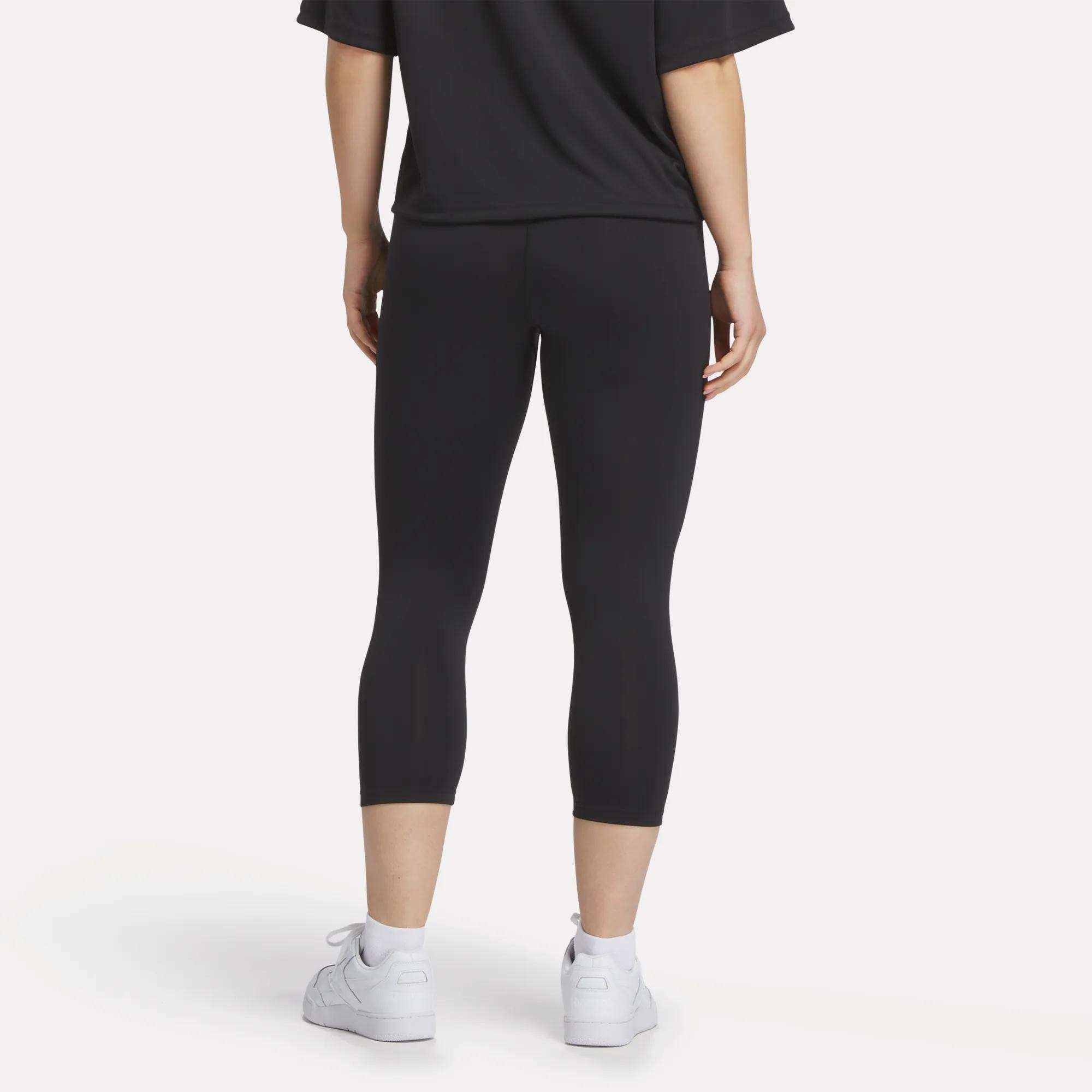 A person stands with their back to the camera, wearing a black t-shirt, white sneakers, and Reebok Women's ID Basketball 3/4 Compression Leggings against a plain light background.