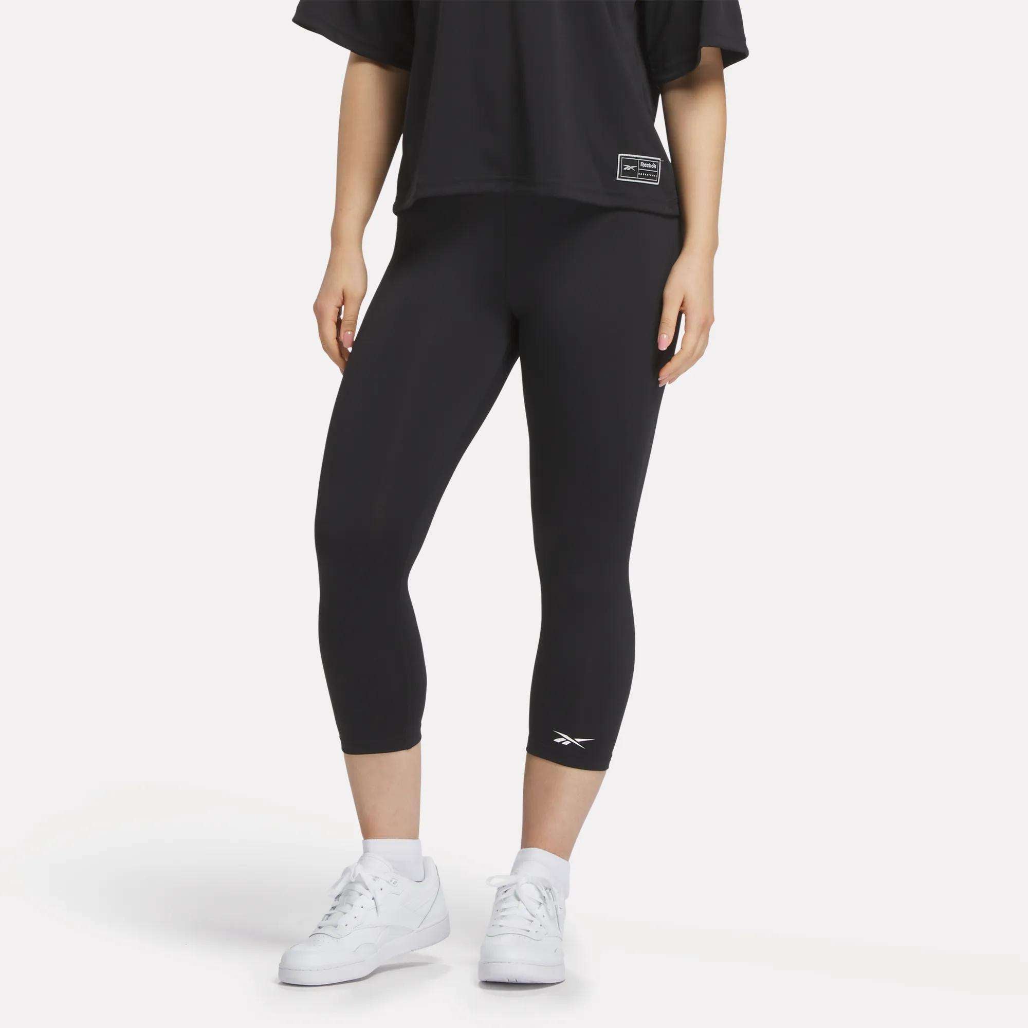 A figure from the shoulders down wears a loose black T-shirt, white sneakers, and Reebok Women's ID Basketball 3/4 Compression Leggings against a plain white background, highlighting the comfort and fit of these leggings.