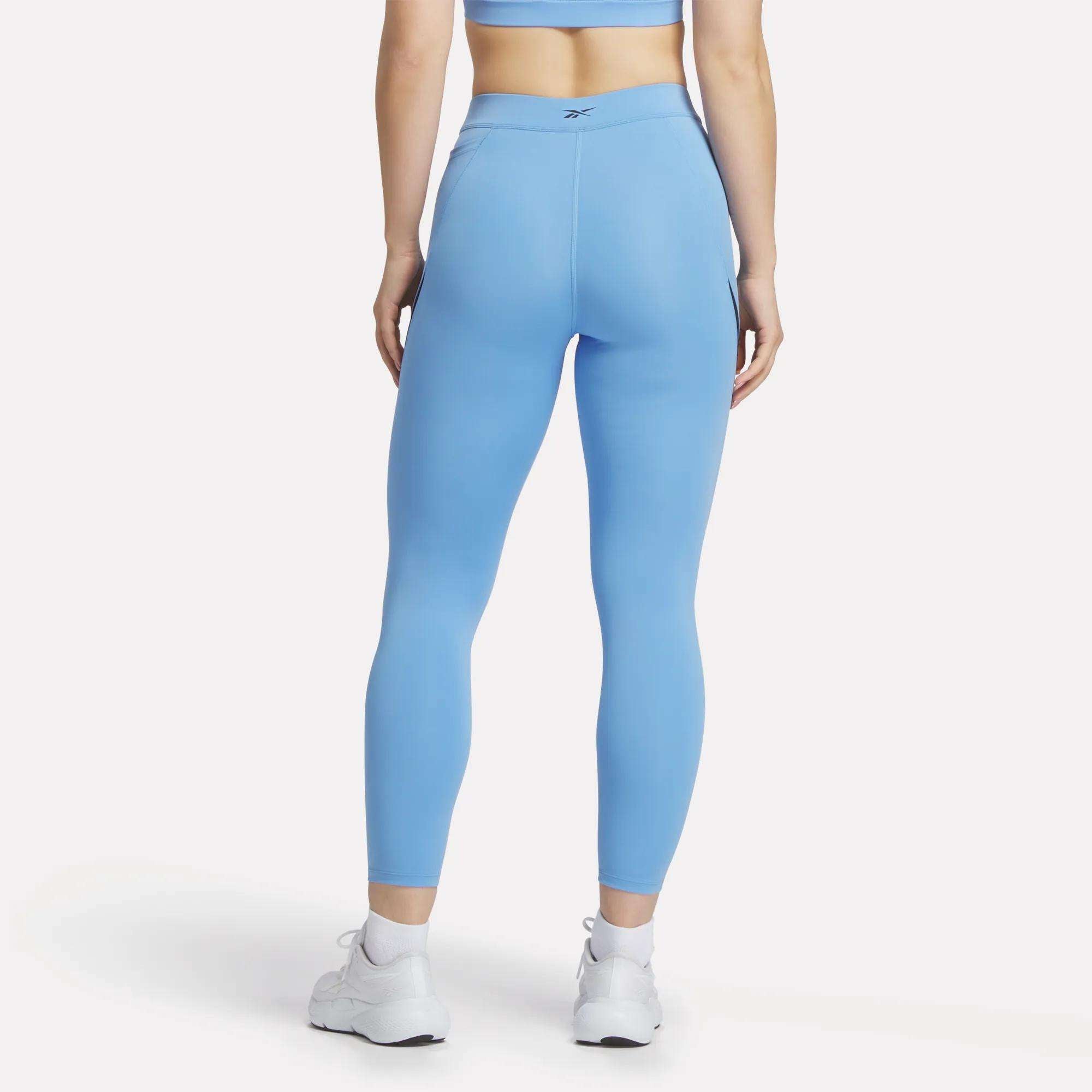 A woman stands facing away in Reebok Women's Lux High-Rise Colorblock Leggings and a light blue sports bra, paired with white athletic shoes. The plain white background highlights the comfort of the sweat-wicking fabric.