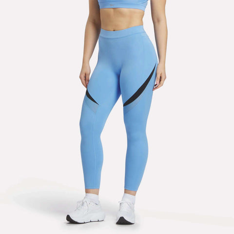 A woman models Reebok Women's Lux High-Rise Colorblock Leggings in light blue with black diagonal stripes, paired with a matching sports bra and white sneakers, standing against a plain white background.