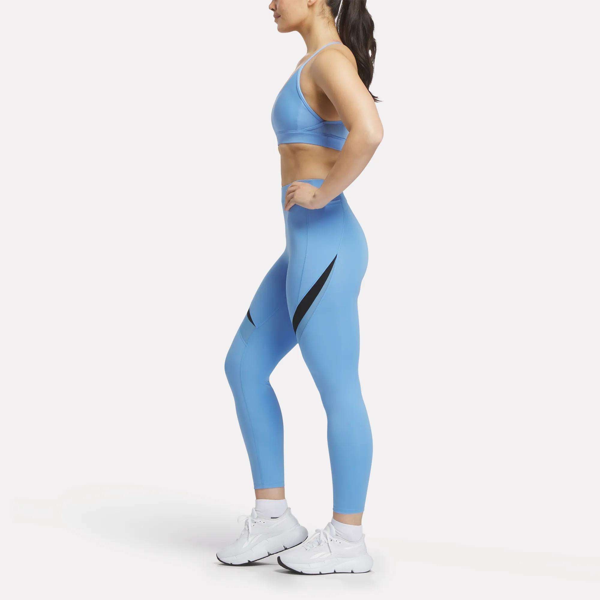 A woman in profile wears Reebok Women's Lux High-Rise Colorblock Leggings in light blue with black accents, paired with a matching sports bra and white athletic shoes, standing against a plain white background.