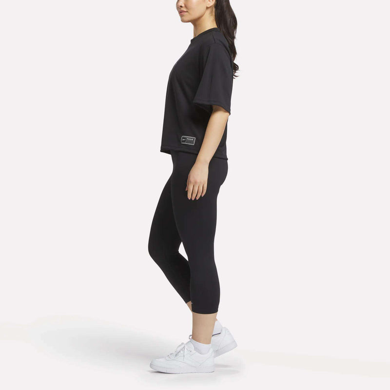 A woman stands in profile wearing a Reebok Women's Basketball Mesh T-Shirt, black cropped leggings, and white sneakers against a plain white background.