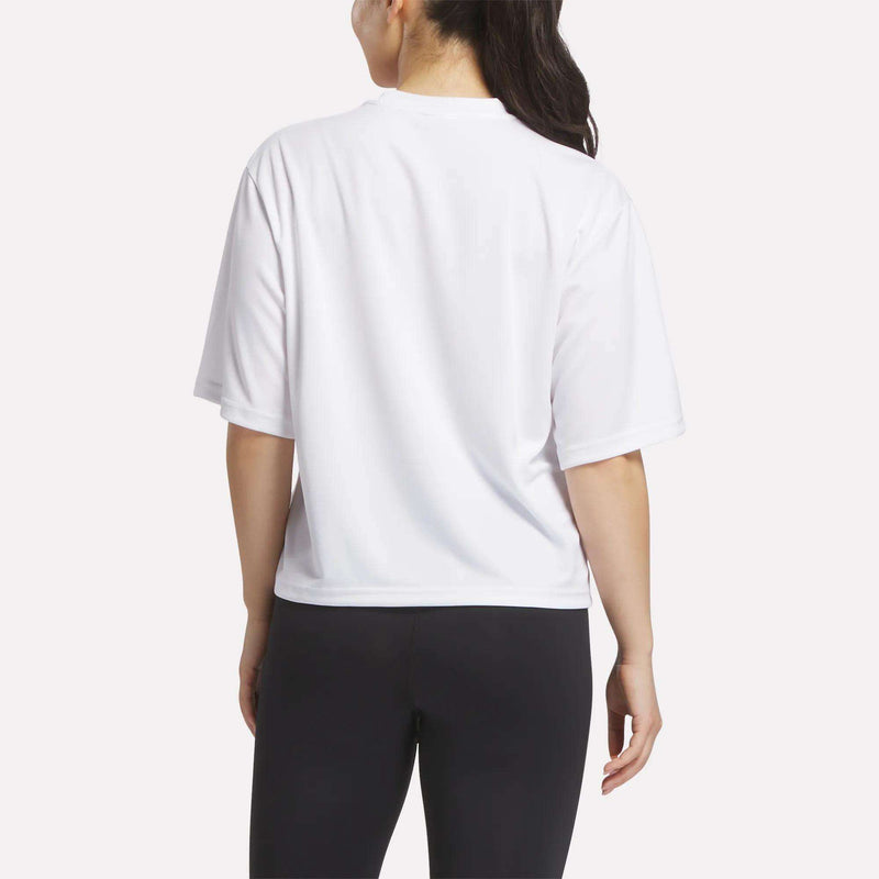 A woman with long dark hair in a ponytail stands with her back to the camera, wearing a loose Reebok Women's Basketball Mesh T-Shirt and black leggings against a plain light background.