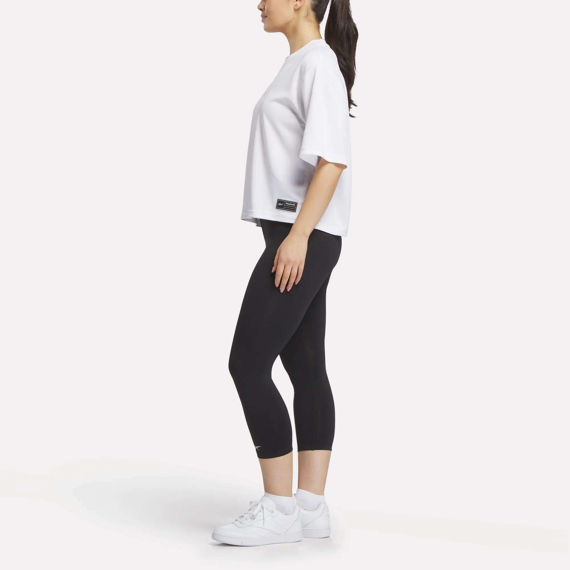 A woman stands sideways in a Reebok Women's Basketball Mesh T-Shirt, black cropped leggings, and white sneakers against a plain white background.