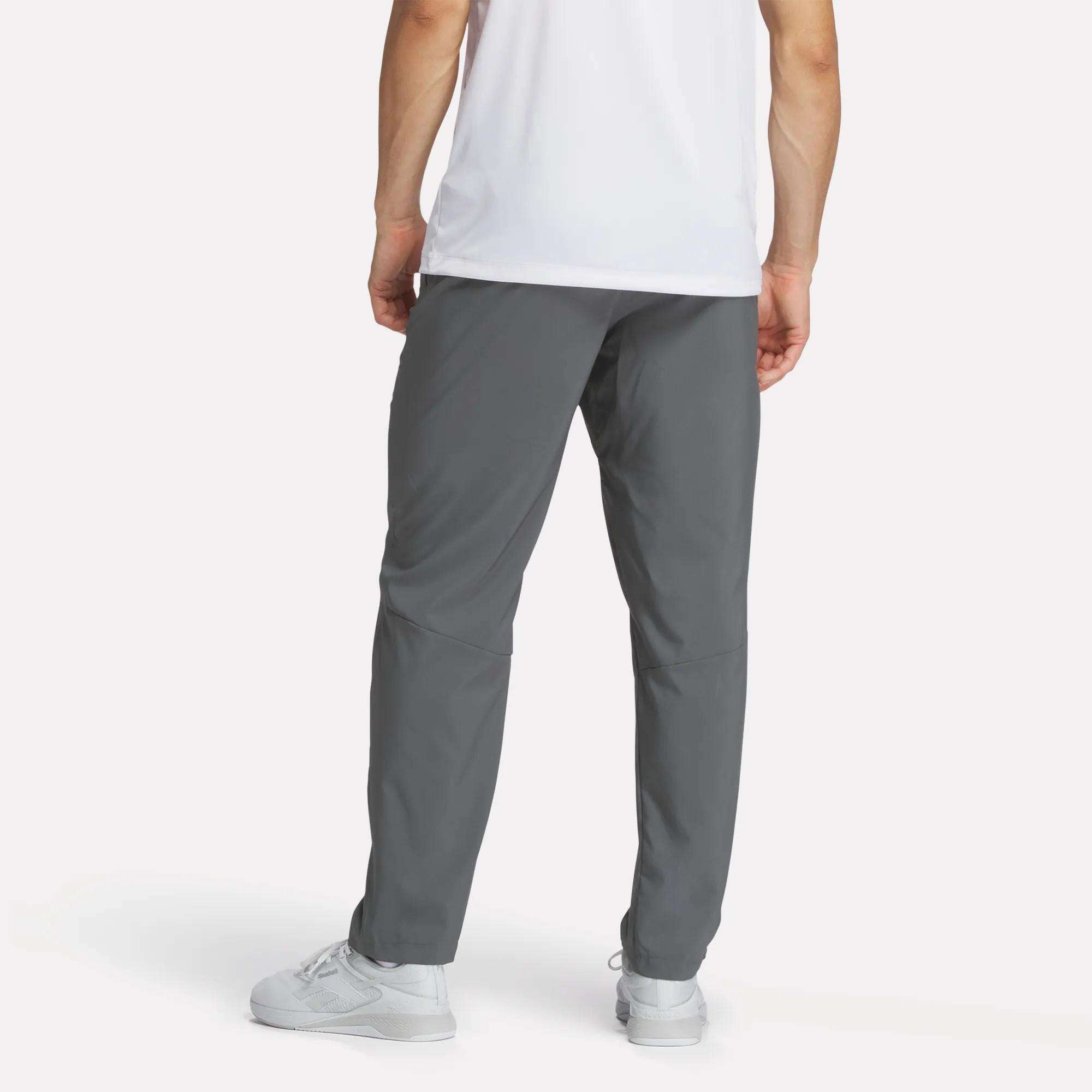 A person models Reebok Men's Woven Pants, paired with a white shirt and sneakers, standing with their back to the camera to display the pants' fit against a plain light background.