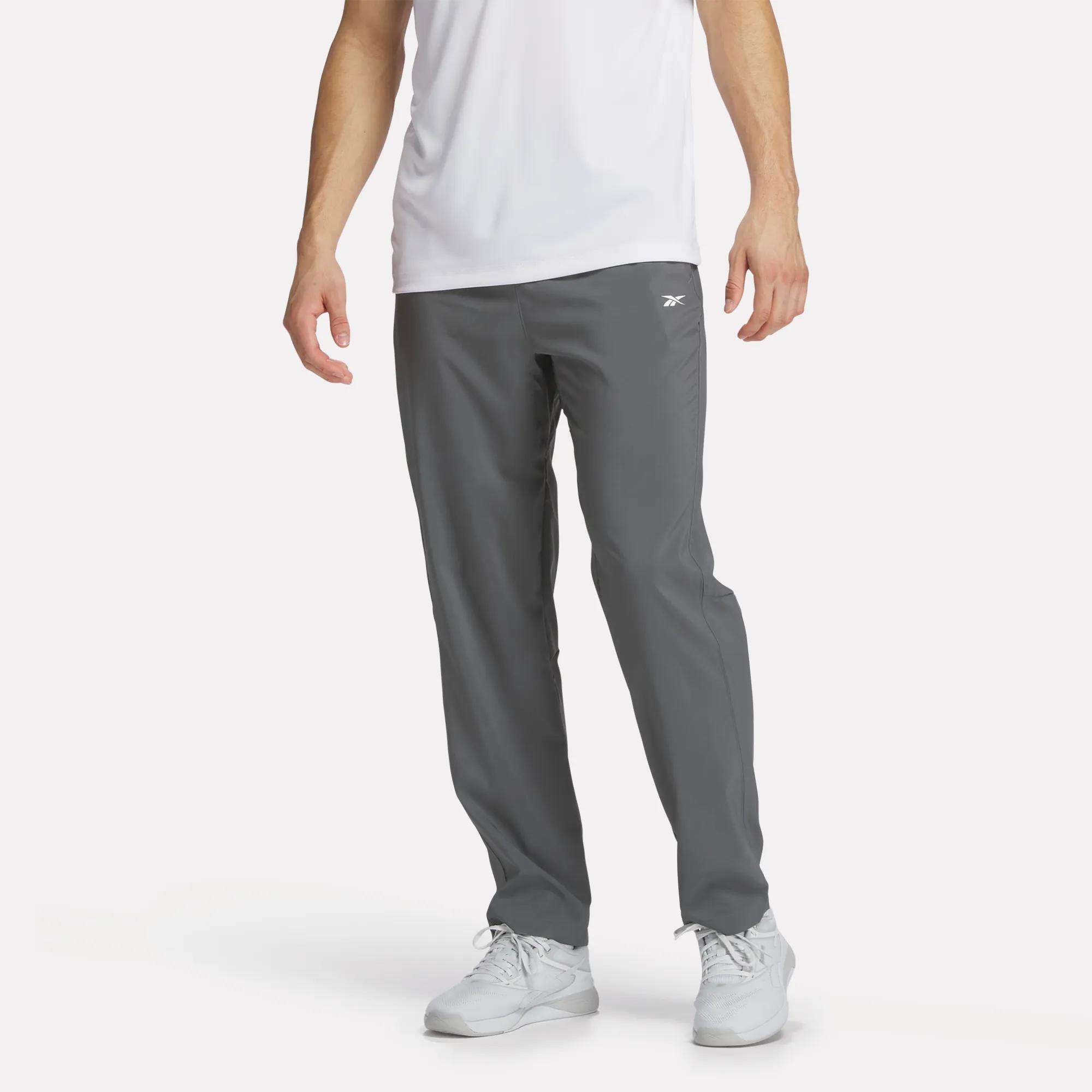 A man wears Reebok Men's Woven Pants in gray with a white tee and sneakers, standing against a plain white background. The focus is on the lightweight polyester pants.