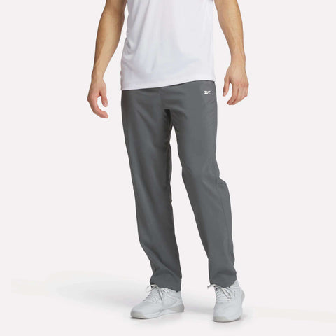 A man wears Reebok Men's Woven Pants in gray with a white tee and sneakers, standing against a plain white background. The focus is on the lightweight polyester pants.