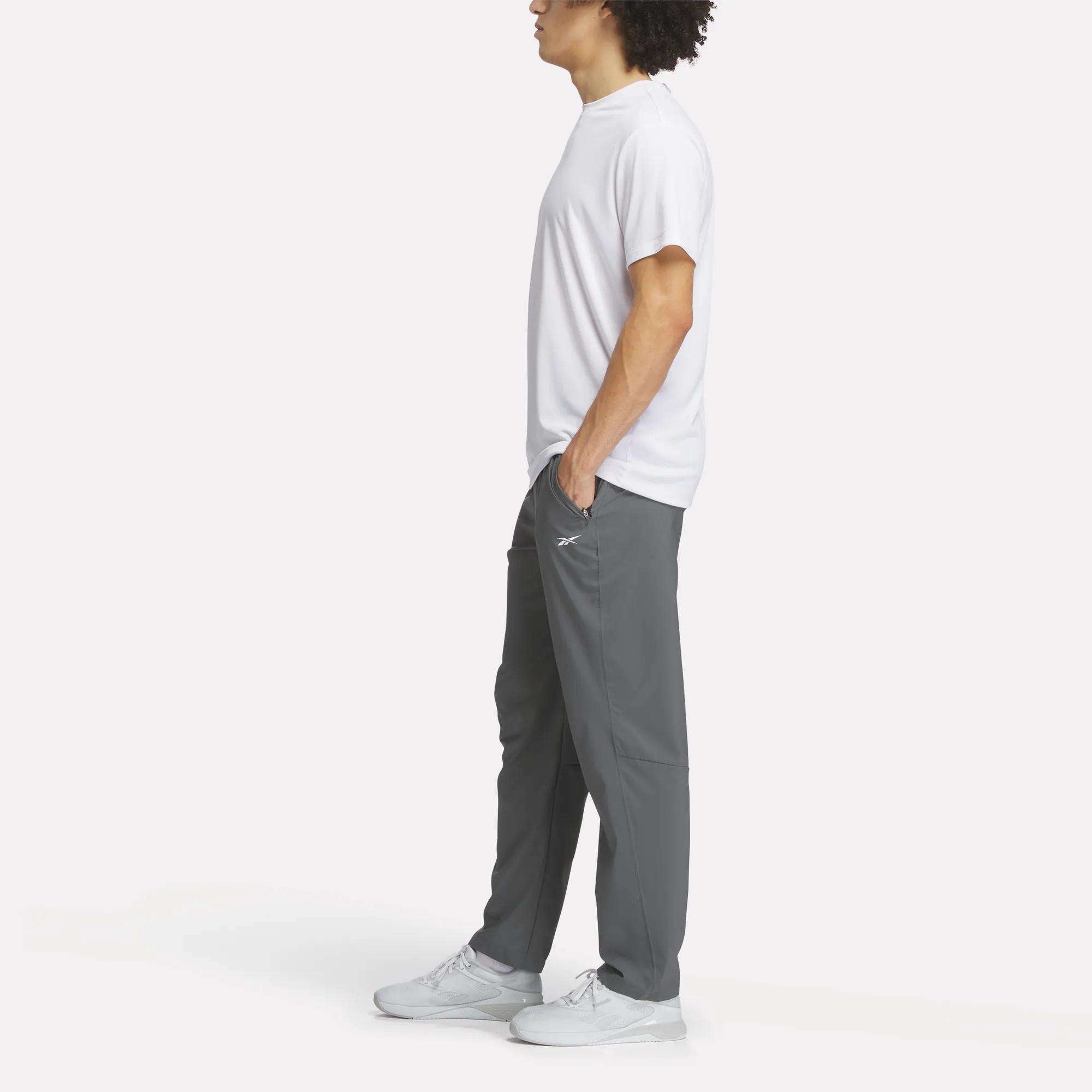 A person stands in profile with hands in pockets, wearing a plain white t-shirt, Reebok Men's Woven Pants, and white sneakers against a light background.