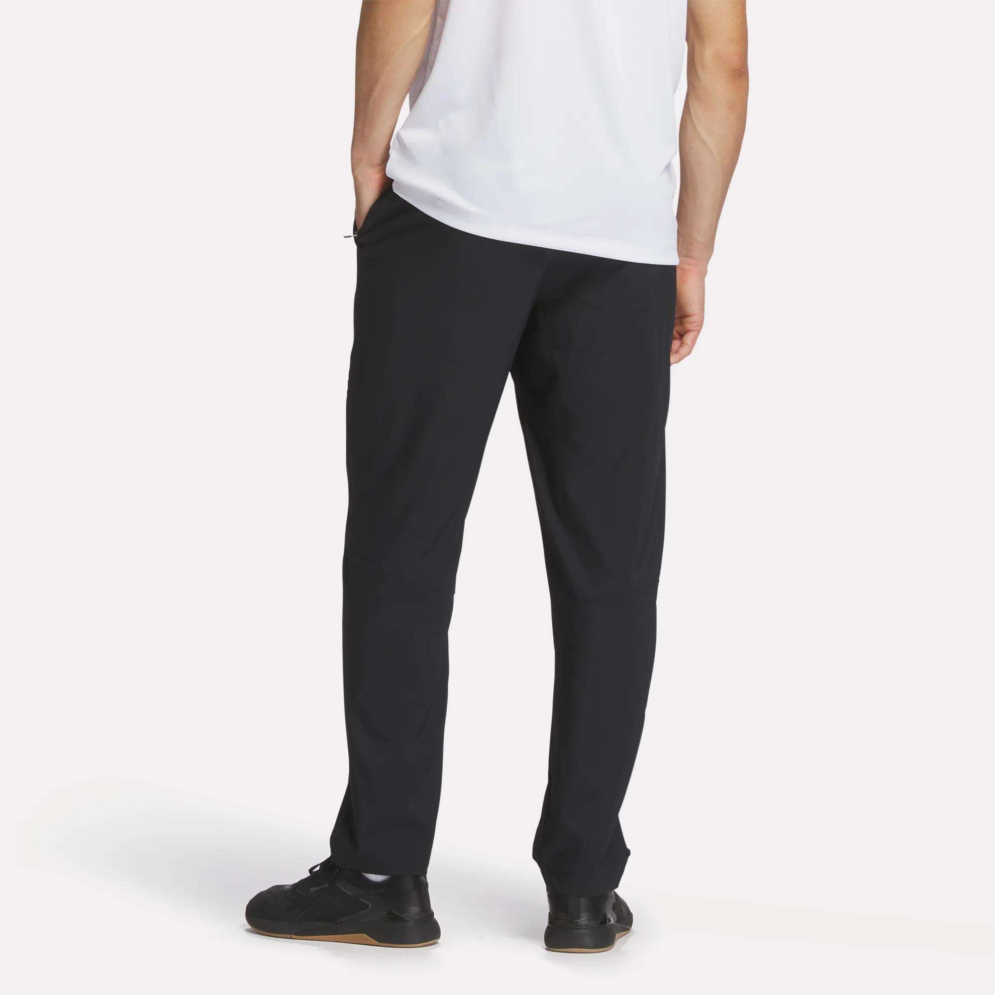 A person stands facing away against a plain white background, wearing black Reebok Men's Woven Pants, a white short-sleeve shirt, and black sneakers.
