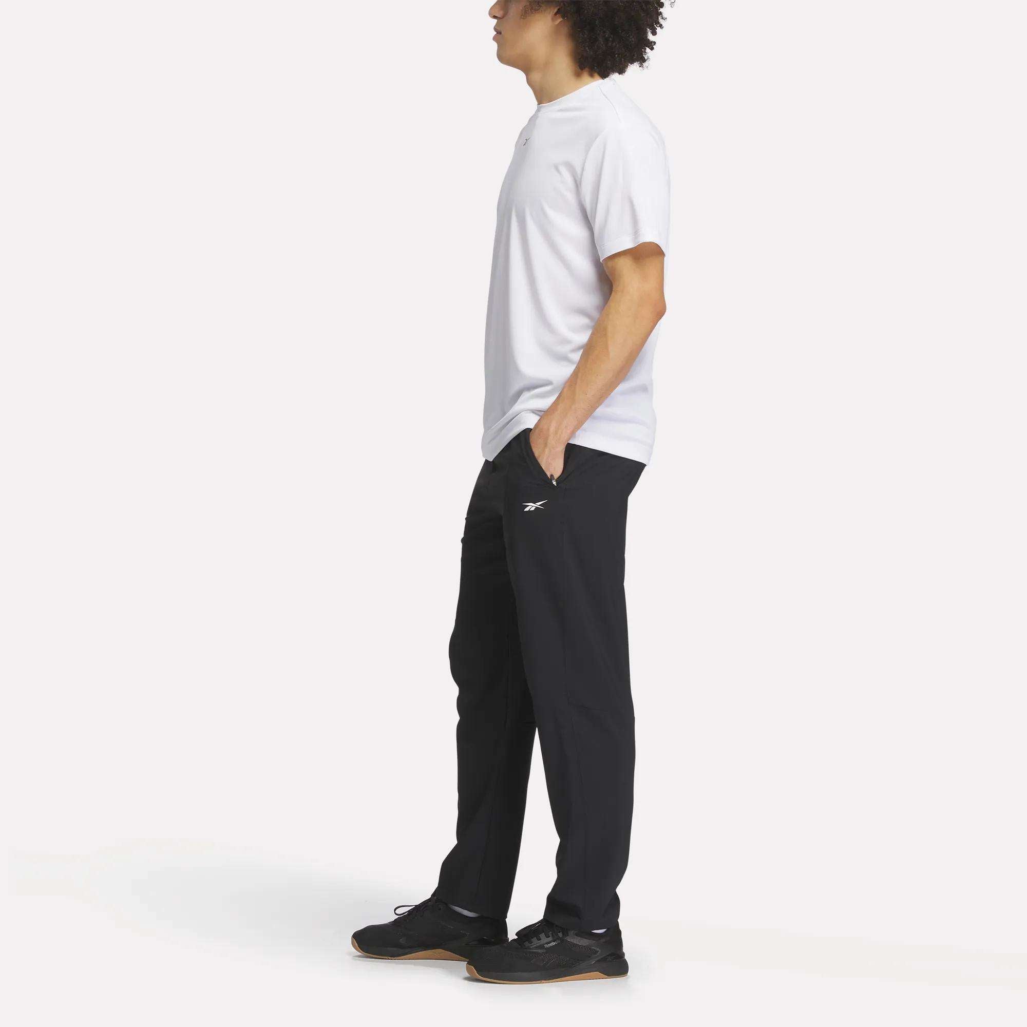 A man stands sideways with hands in pockets, wearing a white T-shirt, Reebok Men's Woven Pants in black, and black sneakers, posed against a plain white background.