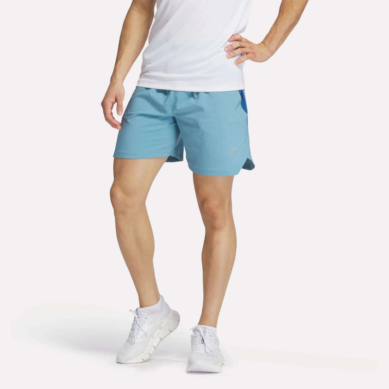 A man stands with one hand on his hip, wearing a white T-shirt, white running shoes, and light blue Reebok Men's Run Shorts with inner tights. His head is not visible, and the background is plain.