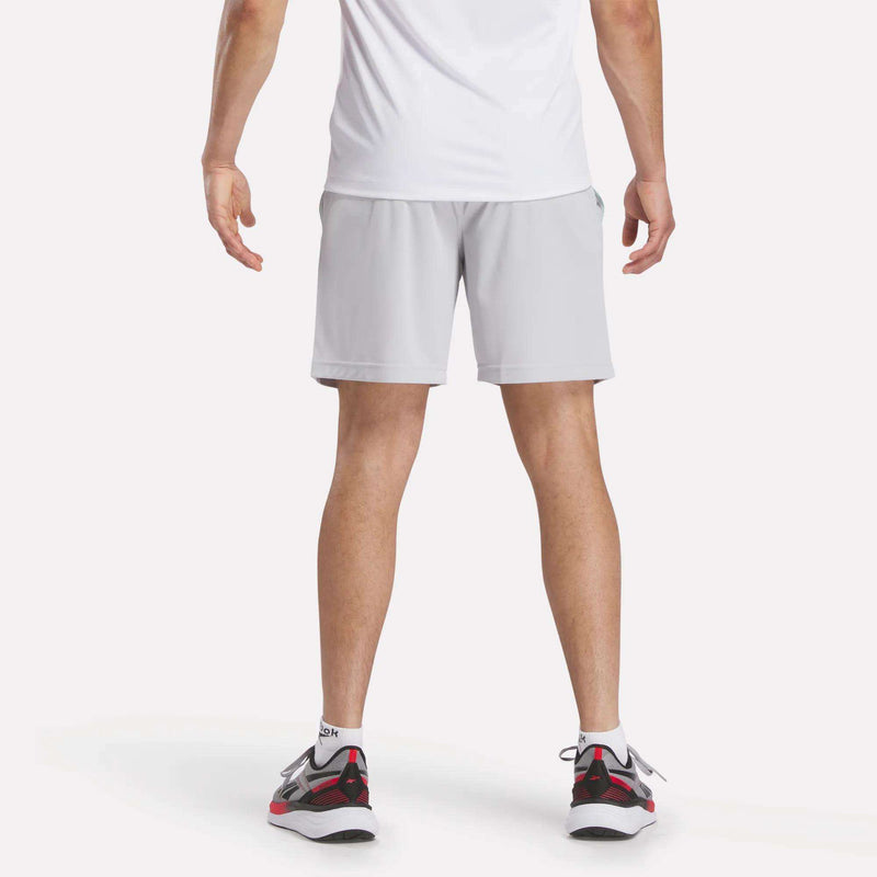 A person wearing a white t-shirt, Reebok Men's Train Blocked Knit Shorts in light gray, and white, black, and red sneakers stands with their back to the camera against a plain white background.