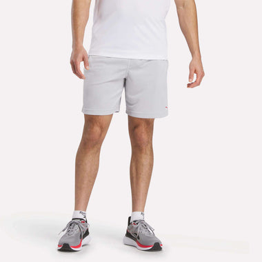 A man wearing a white shirt, Reebok Men's Train Blocked Knit Shorts in light gray, white socks, and gray running shoes with red and black accents stands against a plain white background.