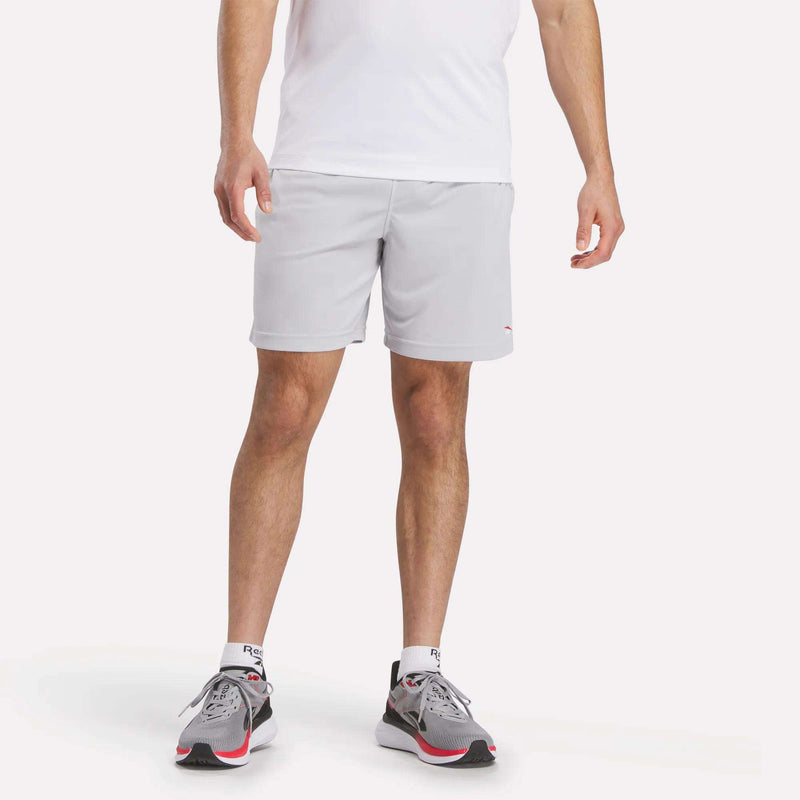 A man wearing a white shirt, Reebok Men's Train Blocked Knit Shorts in light gray, white socks, and gray running shoes with red and black accents stands against a plain white background.