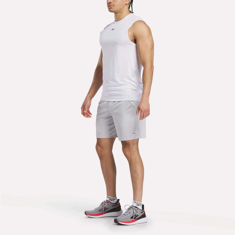 A man stands slightly turned left, wearing a white sleeveless athletic shirt, Reebok Men's Train Blocked Knit Shorts in light gray, and gray athletic shoes with red accents, against a plain white background.