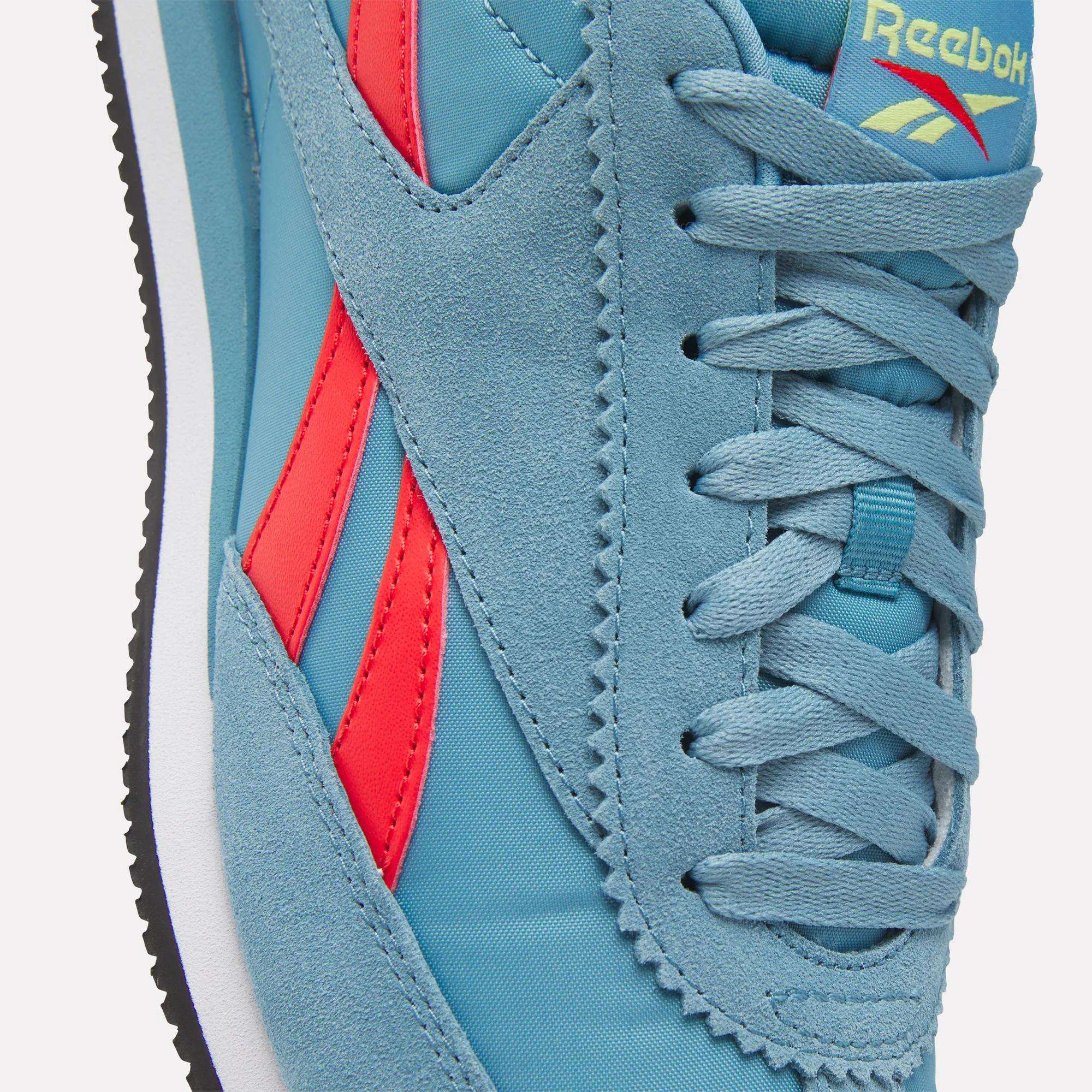 Close-up of Reebok R100 Shoes in blue with red side stripes, featuring a suede and fabric upper, blue laces, and the Reebok logo on the tongue. A black and white low-profile sole completes this '70s running-inspired sneaker.