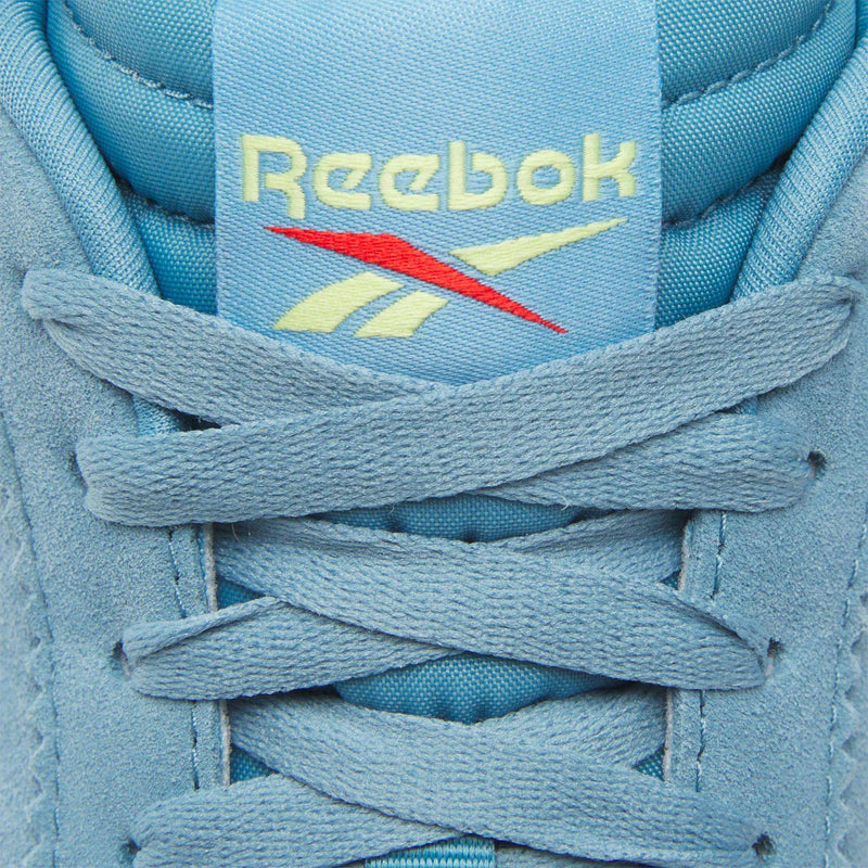 Close-up of the tongue of the Reebok R100 Shoes, featuring light blue laces and an embroidered yellow and red Reebok logo, showcasing their classic '70s running style and low-profile sneaker design.