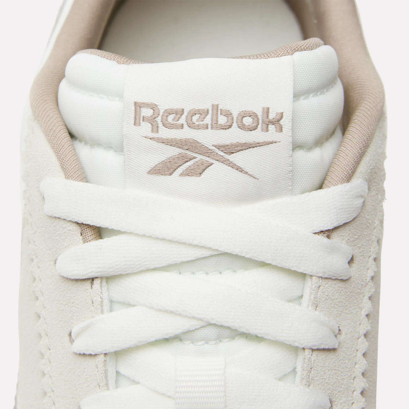 Close-up of white Reebok R100 Shoes, inspired by '70s running style, featuring beige embroidered Reebok logo and name on the tongue, plus white laces threaded through the eyelets.