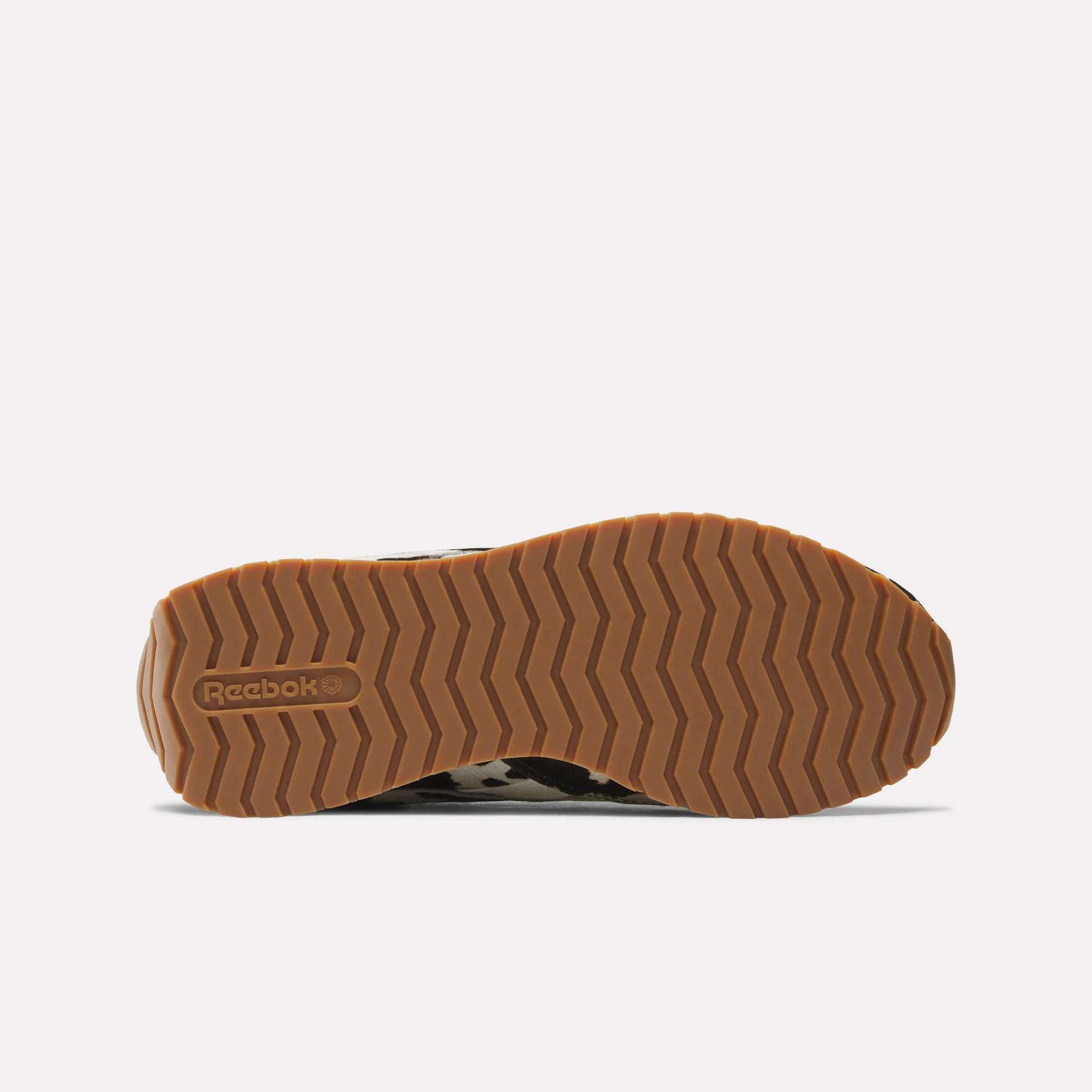 The image shows the bottom of the Reebok Women's Classic AZ Shoes with a tan, zigzag-patterned rubber sole and the embossed Reebok logo near the heel, highlighting their stylish design.