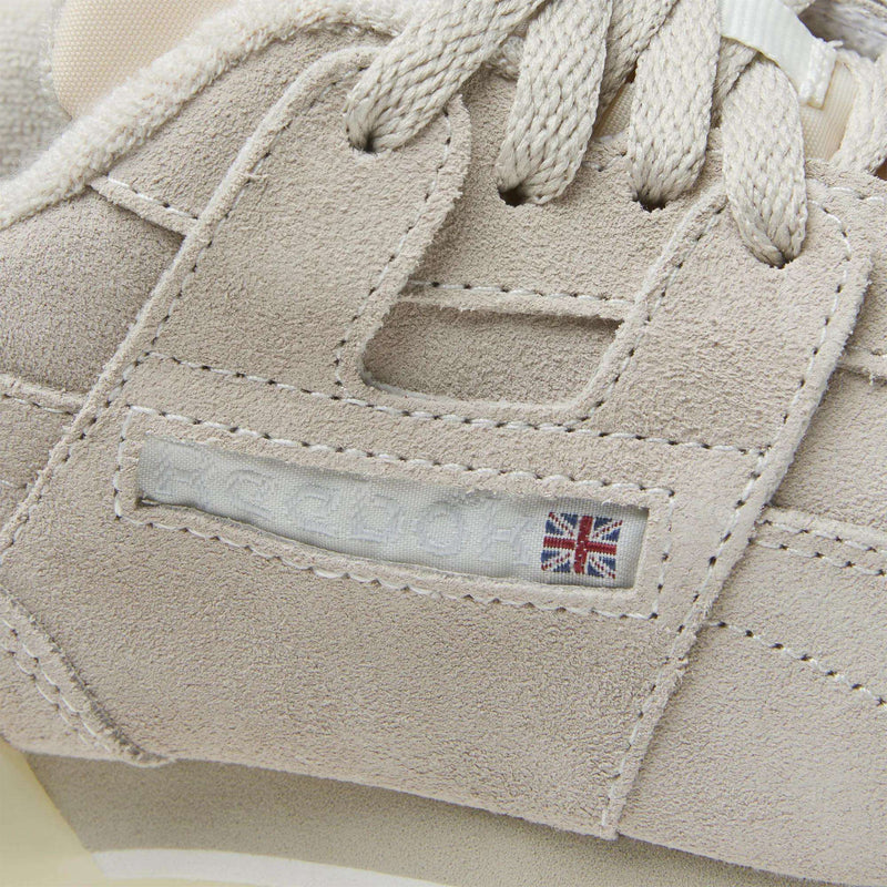 Close-up of Reebok Women's Workout Plus Shoes in beige suede, showcasing stitched details, cream laces, and a small Union Jack flag by the logo—an iconic '80s-inspired style for women's workout footwear.
