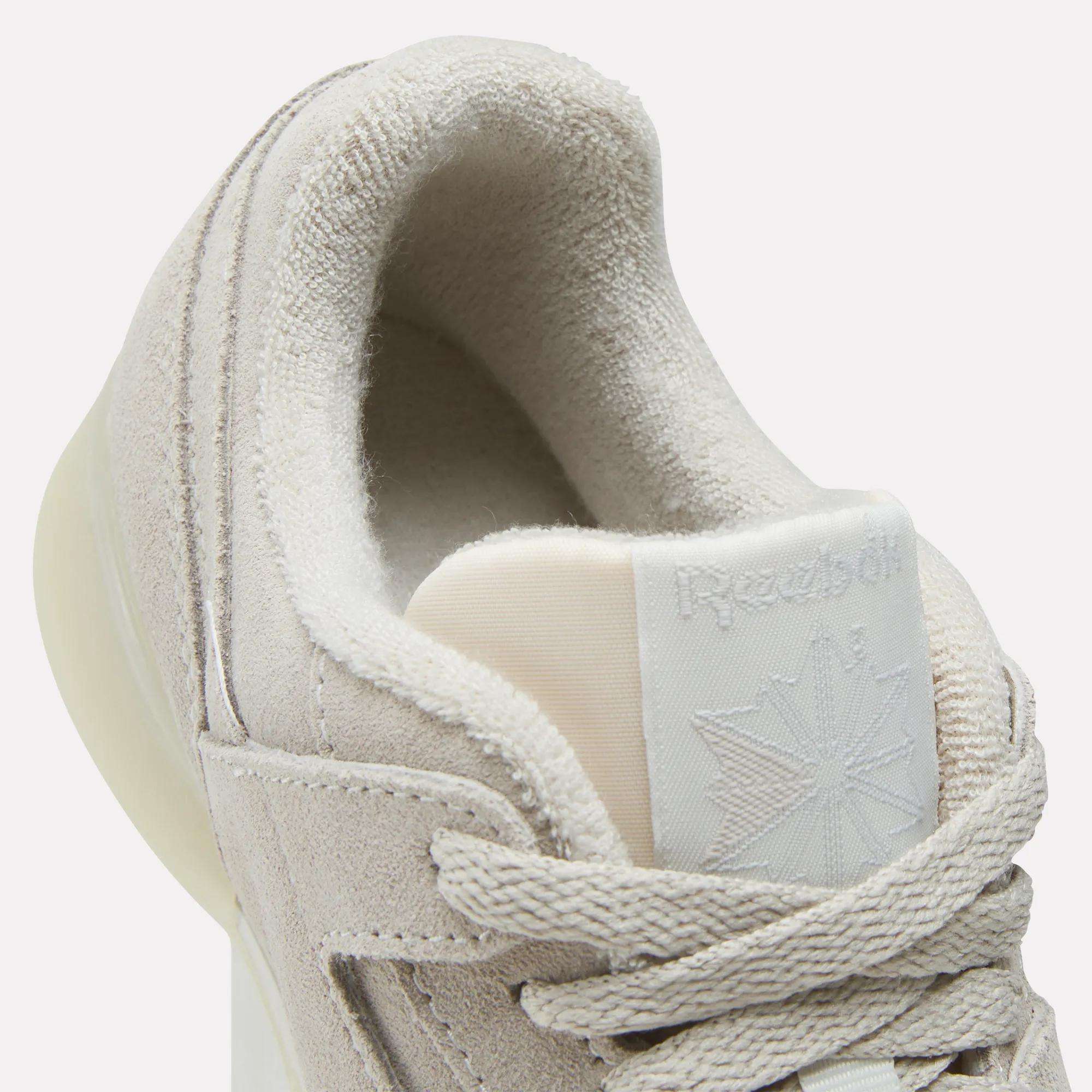 Close-up of Reebok Women's Workout Plus Shoes in light beige, featuring suede texture, thick laces, and a partially visible tongue logo. Plush inner lining adds comfort, capturing an '80s fitness look ideal for women's workout footwear.