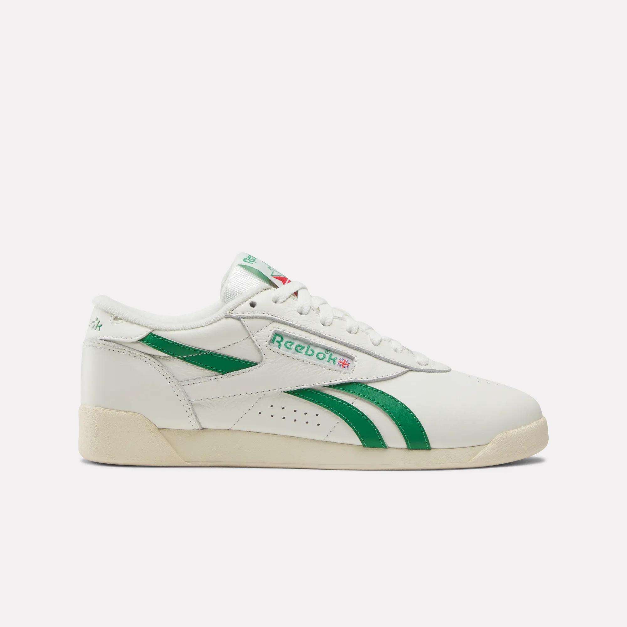 The Reebok Women's Freestyle Lo Shoes feature a white design with green stripes, green logo details, and an off-white EVA midsole, shown in side profile on a plain background.