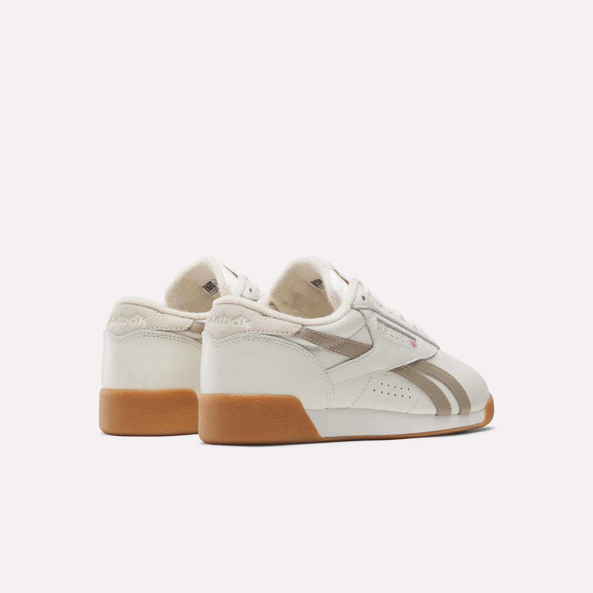 The Reebok Women's Freestyle Lo Shoes in white with beige accents and gum rubber soles are shown from the back and side on a plain white background—perfect retro sneakers for a timeless look.