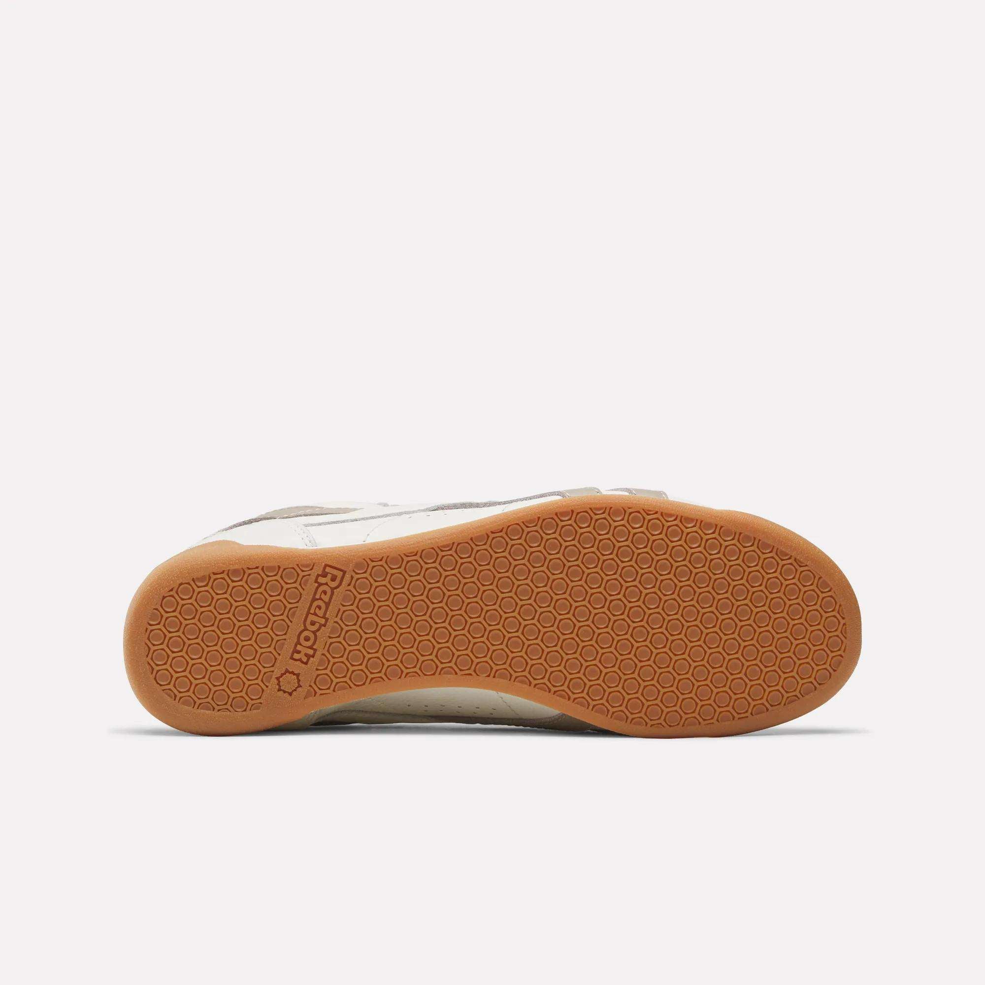 The image shows the bottom view of the Reebok Women's Freestyle Lo Shoes with a light brown, textured rubber sole featuring the Reebok logo near the heel. The partially visible upper is classic white, reflecting a retro sneaker style.