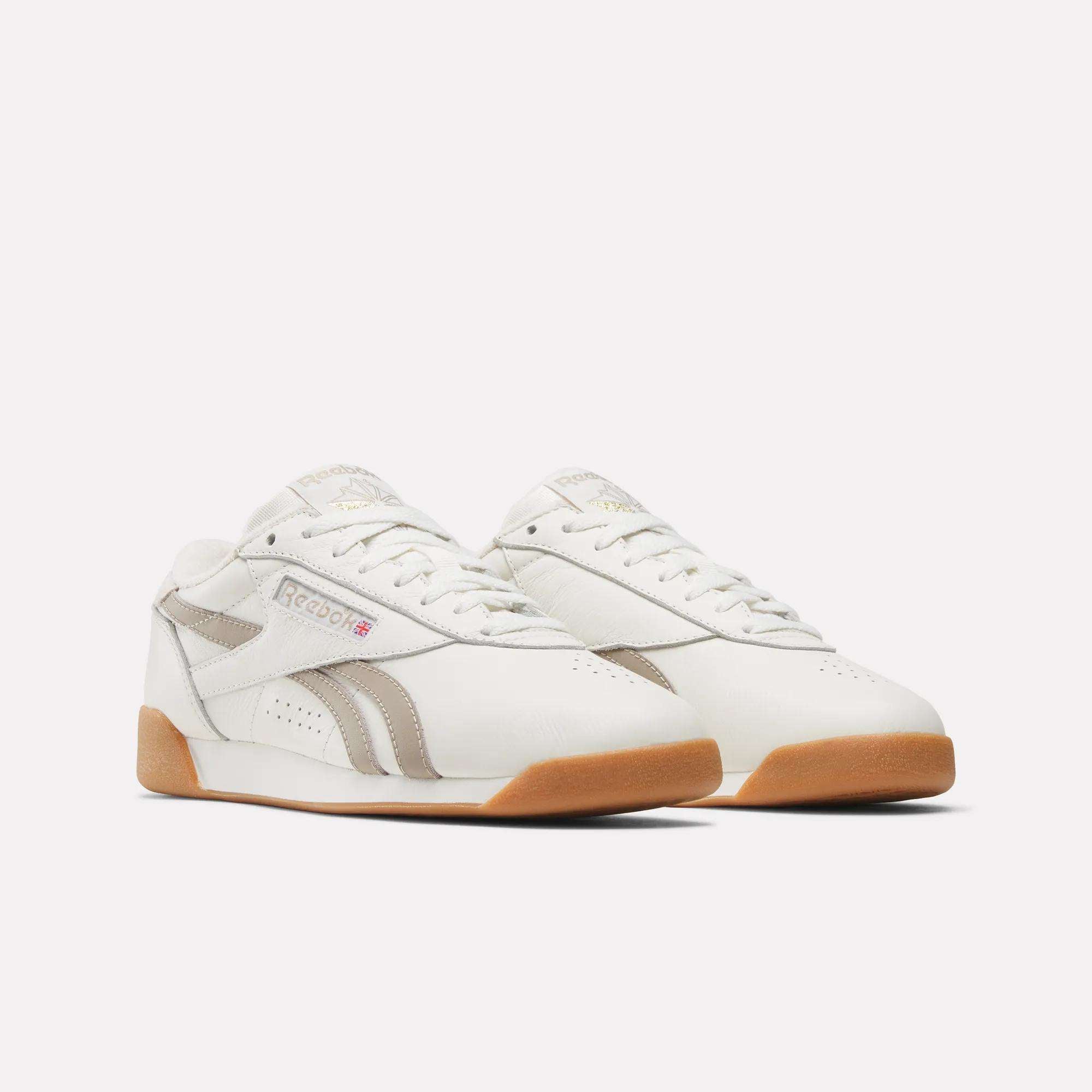The Reebok Women's Freestyle Lo Shoes feature a white design with light beige accents, gum soles, and the iconic Reebok logo on the side and tongue, showcasing a classic retro sneaker style against a plain white background.