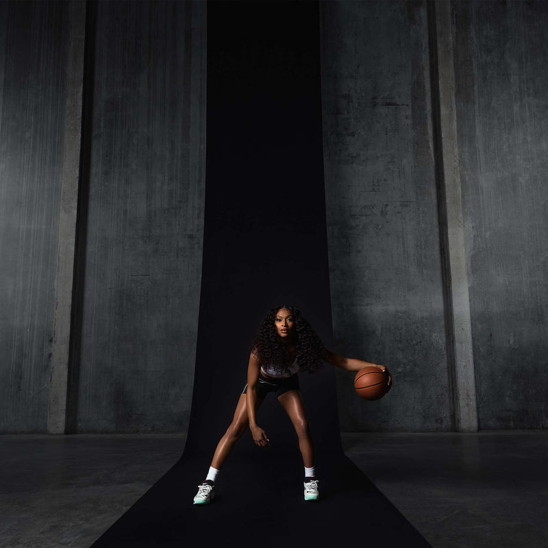 A woman with long curly hair dribbles a basketball on a black runway in front of gray concrete walls, wearing a sports outfit and Reebok Engine A 26 Basketball Shoes designed for energy return and stability, appearing focused and athletic.