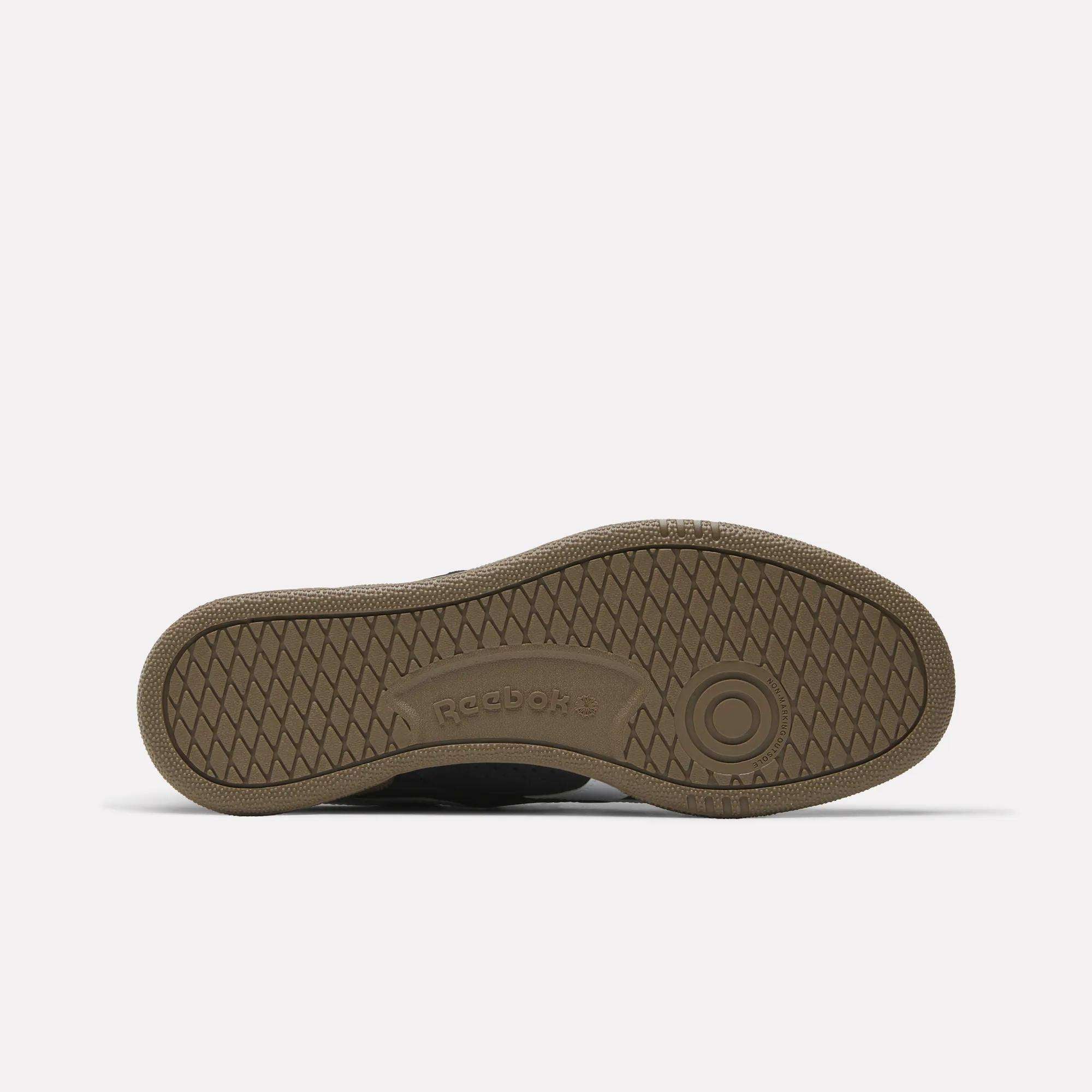The image shows the sole of the Reebok Club C Grounds UK Shoes, featuring a brown, textured non-slip pattern with "Reebok" embossed near the heel and a circular design at the ball, set against a plain background.