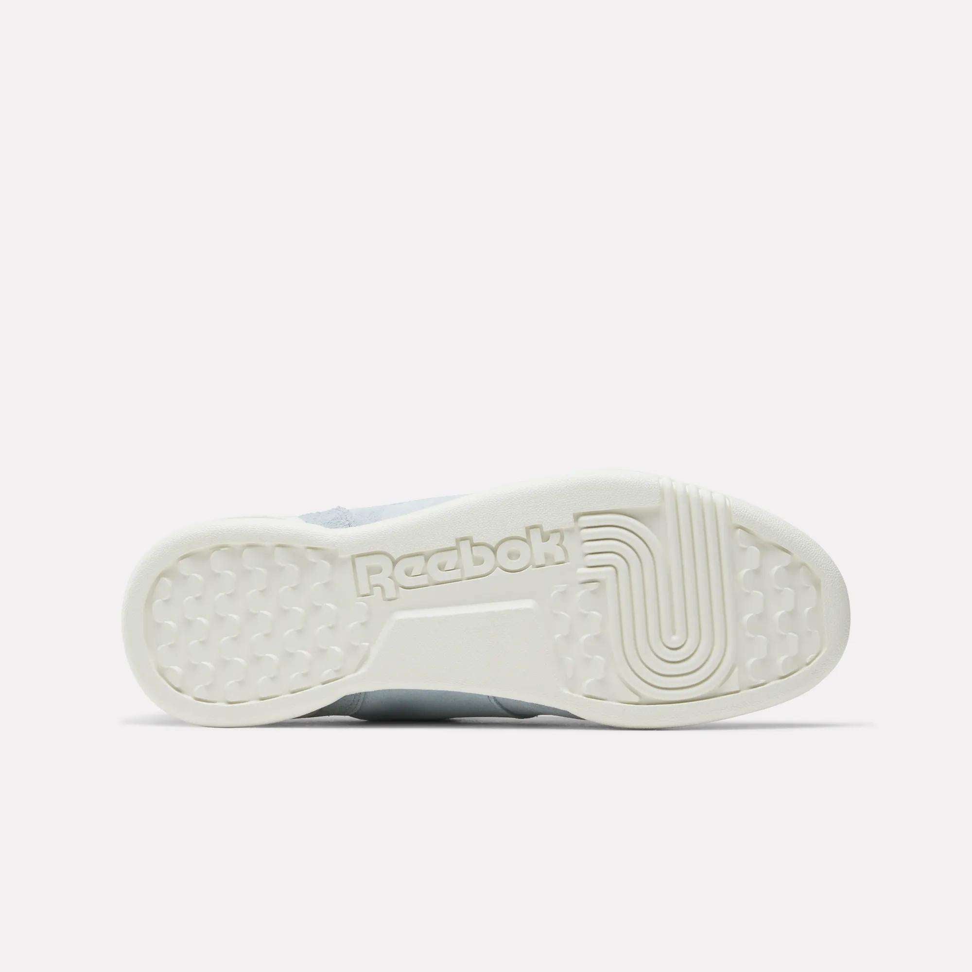 The image shows the white rubber sole of the Reebok Women's Workout Plus Shoes, viewed from below. The sole features the Reebok logo and a mix of hexagonal and linear patterns, reflecting classic '80s fitness style.