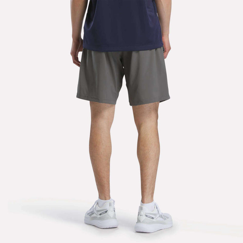 A person faces away, wearing a navy shirt, Reebok Men's Woven Shorts in gray, and white sneakers against a plain background.